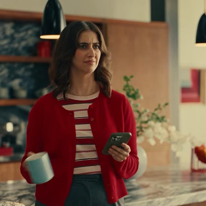 A woman in a red cardigan holding a mug and phone in a modern kitchen