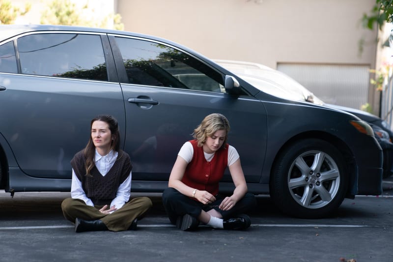 Two characters sitting on the asphalt beside a parked car