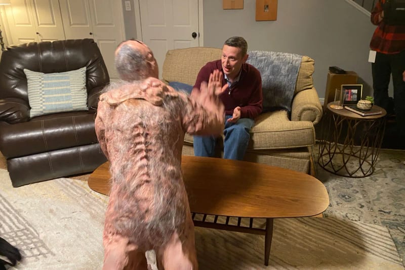 A hairy, prosthetic monster character confronting a man on a couch