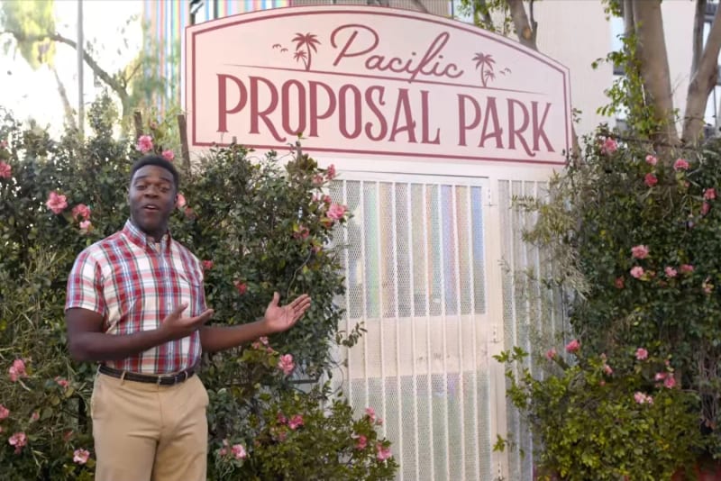 A man gesturing beside the Pacific Proposal Park sign, set dressed with flowers