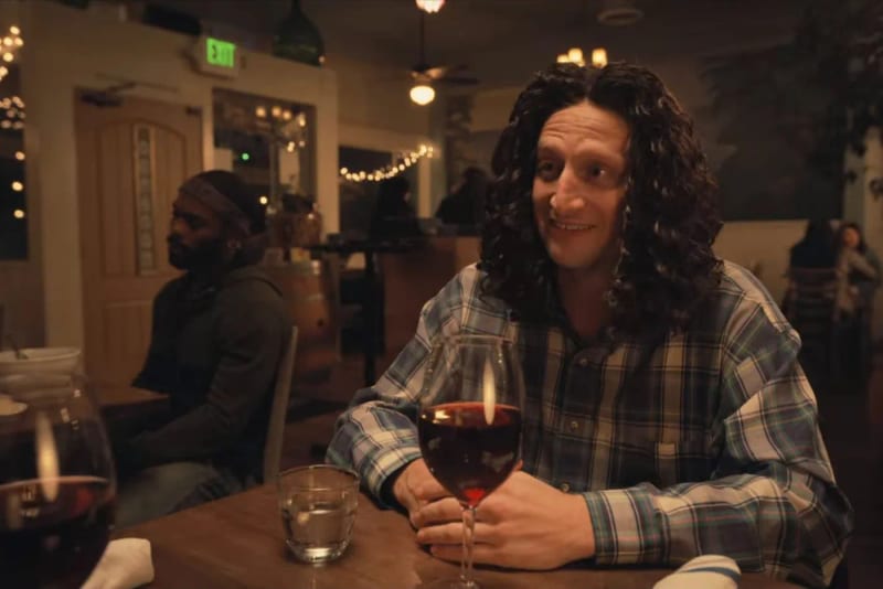 A man with a long curly wig seated at a restaurant table with wine