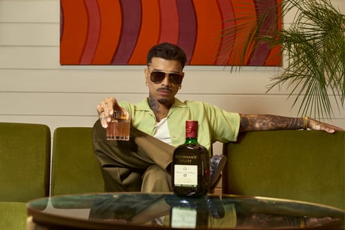 A man sitting on a green couch with a glass and bottle of Buchanan’s Deluxe
