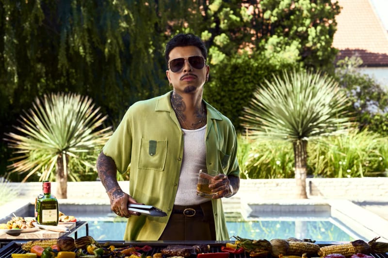 A man grilling by a pool holding a drink, with Buchanan’s Deluxe on the table
