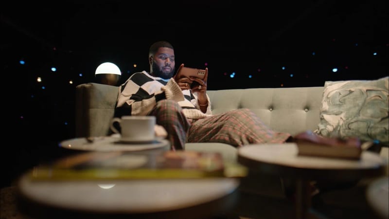A man relaxing on a couch checking his phone with string lights in the background