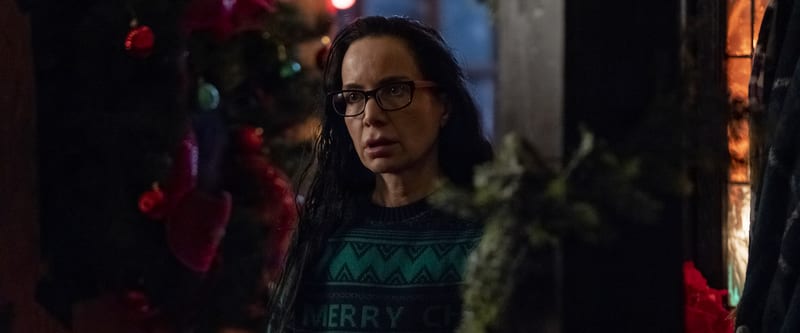 A woman in glasses looking startled beside a lit Christmas tree