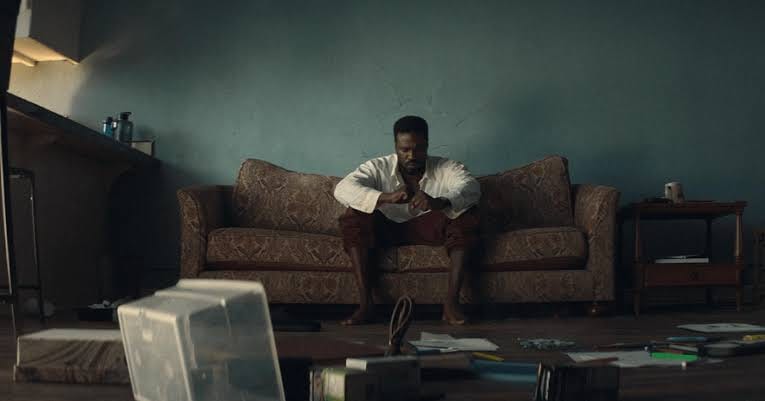 Man sitting on a patterned couch in a cluttered apartment